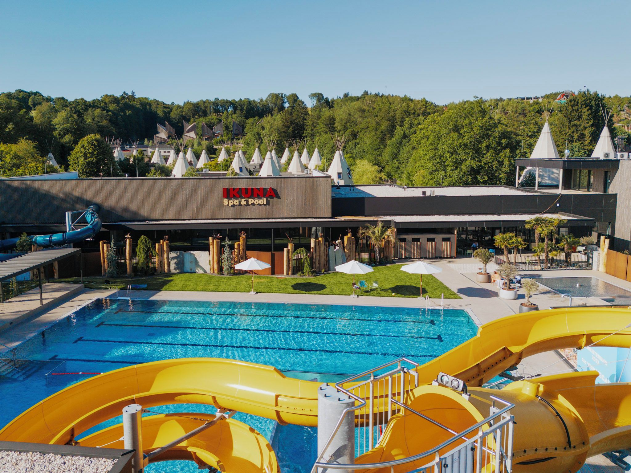 Ein Schwimmbecken mit Wasserrutschen, ein Spa- und Poolgebäude sowie eine begrünte Anlage mit Bäumen und Sonnenschirmen.
