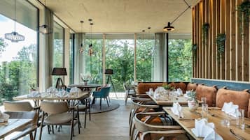 A restaurant with multiple dining tables and chairs, large windows offering a view of greenery, and hanging pendant lights.