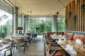 Un restaurant doté de nombreuses tables et chaises, de grandes fenêtres donnant sur un paysage verdoyant et de luminaires suspendus.