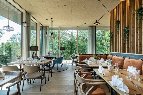 A restaurant with multiple dining tables and chairs, large windows offering a view of greenery, and hanging pendant lights. - Ikuna Naturresort (Natternbach)
