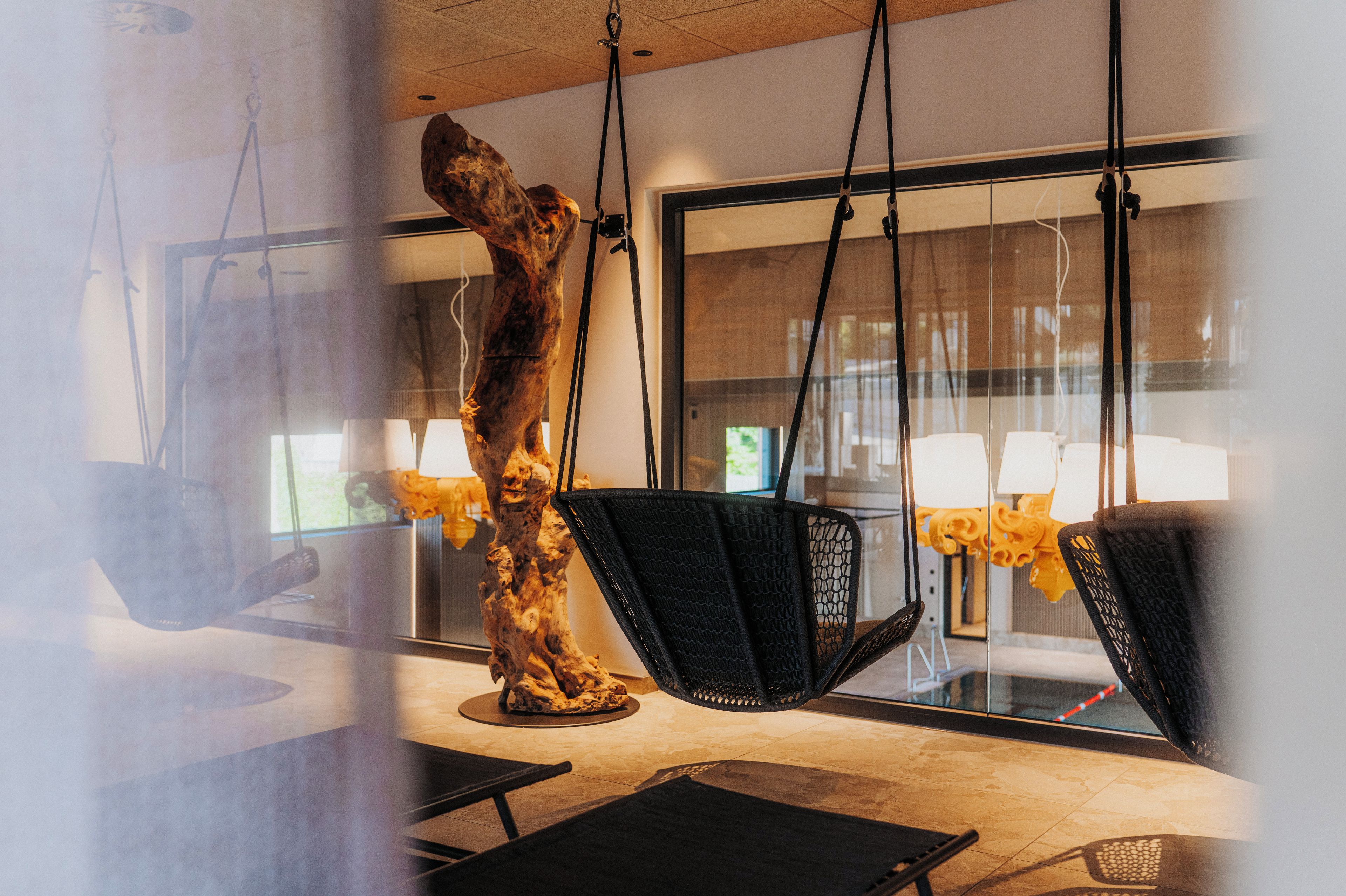 Ein modernes Interieur mit einem großen Fenster, einer hängenden Holzskulptur und schwarzen Stühlen mit strukturierter Oberfläche.