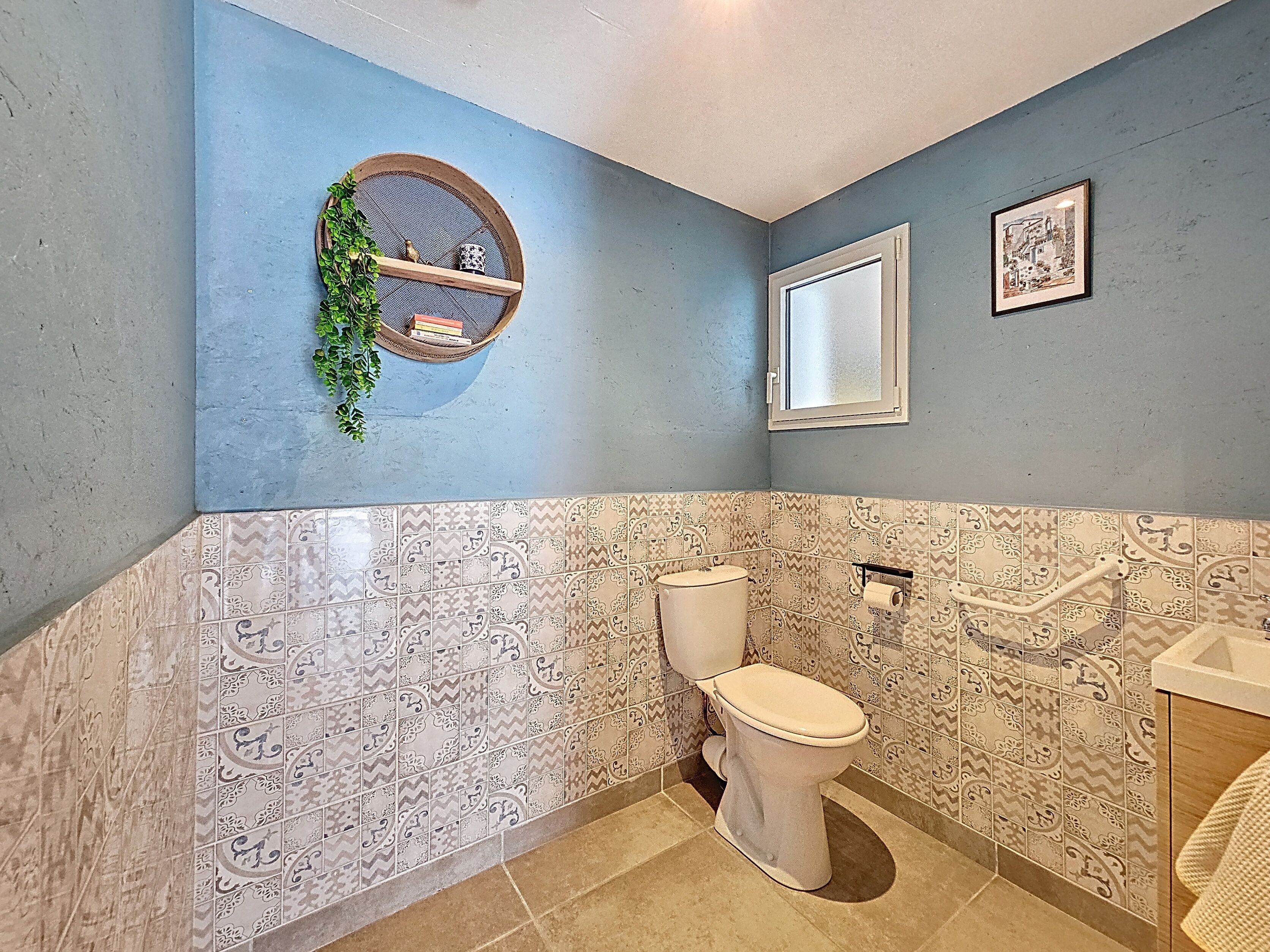 Une salle de bain avec des toilettes, des carreaux décoratifs, une étagère avec une plante et un tableau encadré au mur.