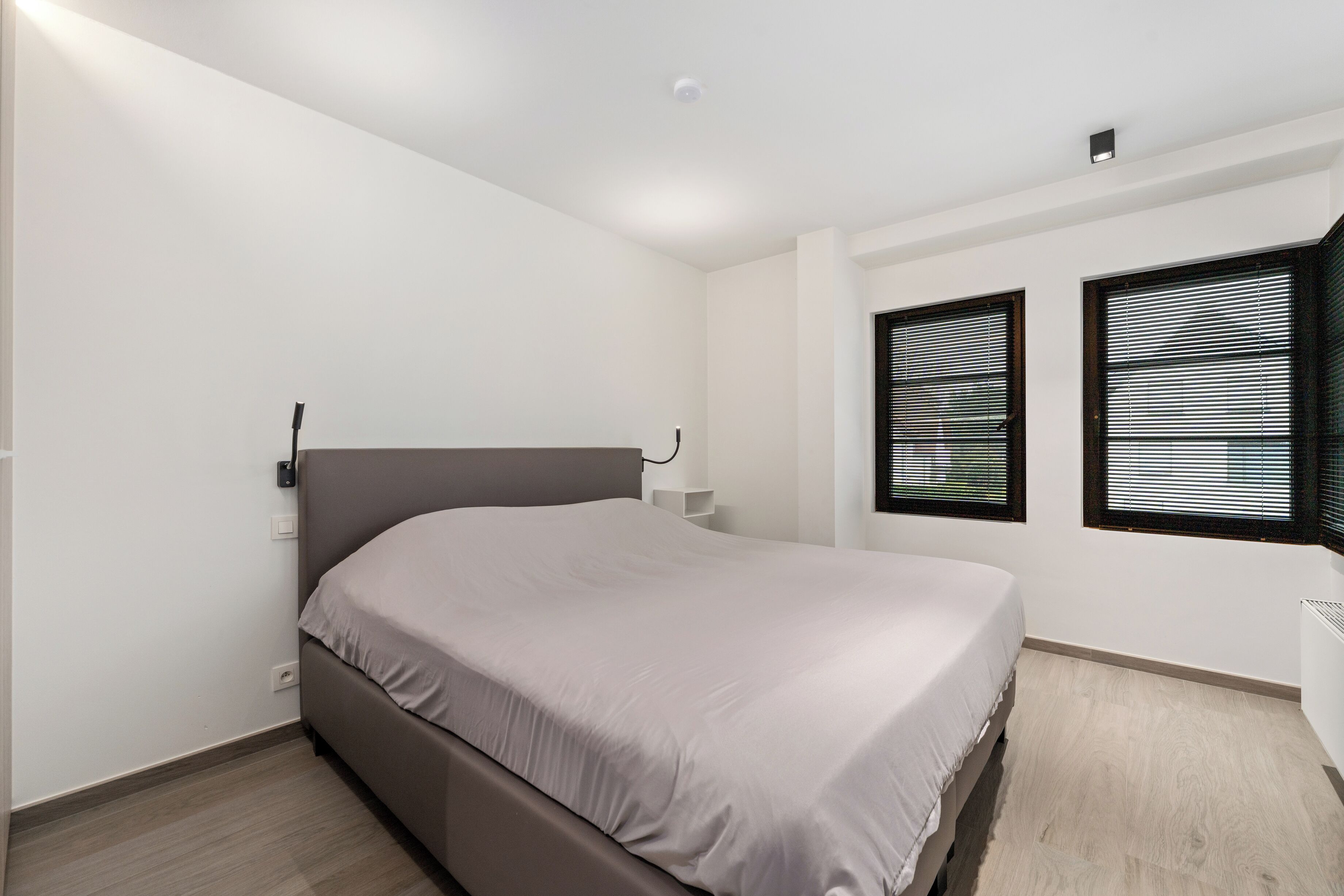 Ein Schlafzimmer mit einem Einzelbett, weißen Wänden, zwei Fenstern mit Jalousien, einem Nachttisch und einer Deckenleuchte.