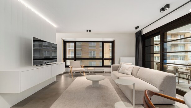 Een moderne woonkamer met een flatscreen televisie, een bank, een salontafel en een fauteuil. Er is een groot raam met zonwering en uitzicht op gebouwen.