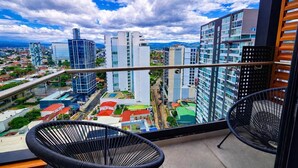 Un balcón con dos sillas de mimbre negro, vistas a un paisaje urbano con edificios modernos y cielo despejado.