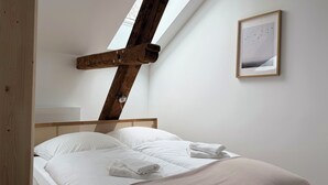 Een slaapkamer met een schuine plafond, een houten hoofdbord, een bed met witte lakens, en een ingelijst schilderij aan de muur.