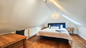 A bedroom with a sloped ceiling, a wooden bed frame, a bedside table, a nightstand, and a window providing natural light. - Y Stabl - 1 Bedroom Apartment - Llanrhidian (Swansea)