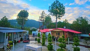 A rural guesthouse with a red-roofed building, a blue-roofed structure, and a paved courtyard with neatly trimmed bushes. - The Chail Heights Hotel & Resort (Shimla)