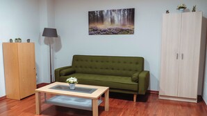 Een kamer met een groene bank, een houten kast, een vloerlamp en een schilderij van een bos.