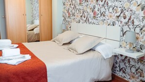 Een net opgemaakt bed met een rode deken, twee kussens en een wit hoofdbord. Er staat een klein nachtkastje bij, met een lamp en een spiegel aan de muur.