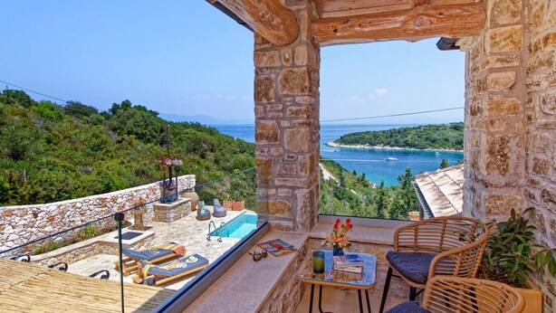 A balcony with a stone wall, a pool, wicker furniture, and a view of a coastal landscape. - Serenity Near the Sea (Gaios)