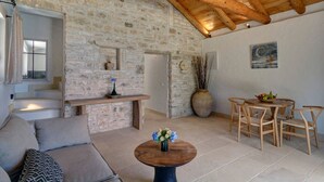 A spacious living area with a stone wall, wooden ceiling beams, a sofa, a round table with flowers, and a dining set. - Serenity Near the Sea (Gaios)