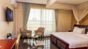 A hotel room with a bed, two armchairs, a small table, a TV, and a window with curtains. - Nican Resort Hotel (Kampala)