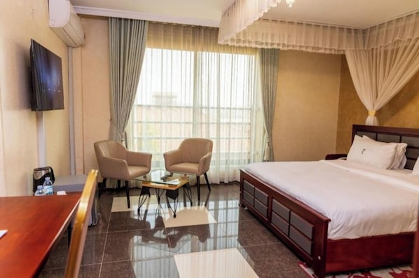 A hotel room with a bed, two armchairs, a small table, a TV, and a window with curtains. - Nican Resort Hotel (Kampala)