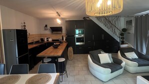 A modern kitchen with a central island, dark cabinetry, and a microwave. There is a dining area with a table and chairs, and a living area with a sofa and a unique ceiling light fixture.