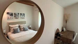 A bedroom with a round mirror, a bed with pillows, a nightstand, a table with a surfboard, and wall art.