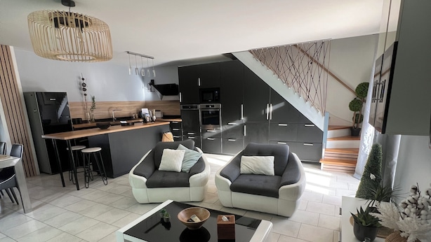 A modern living room with a glass coffee table, two sofas, a dining area with a kitchen in the background, and a staircase leading to an upper level.