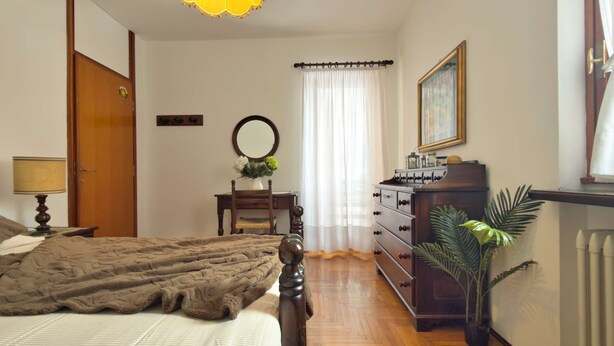 Quarto com cama, penteadeira, espelho, abajur, planta e janela com cortinas.