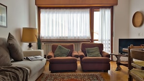 Uma sala de estar acolhedora com sofá, duas poltronas, uma mesa de centro, televisão e uma janela grande com cortinas.