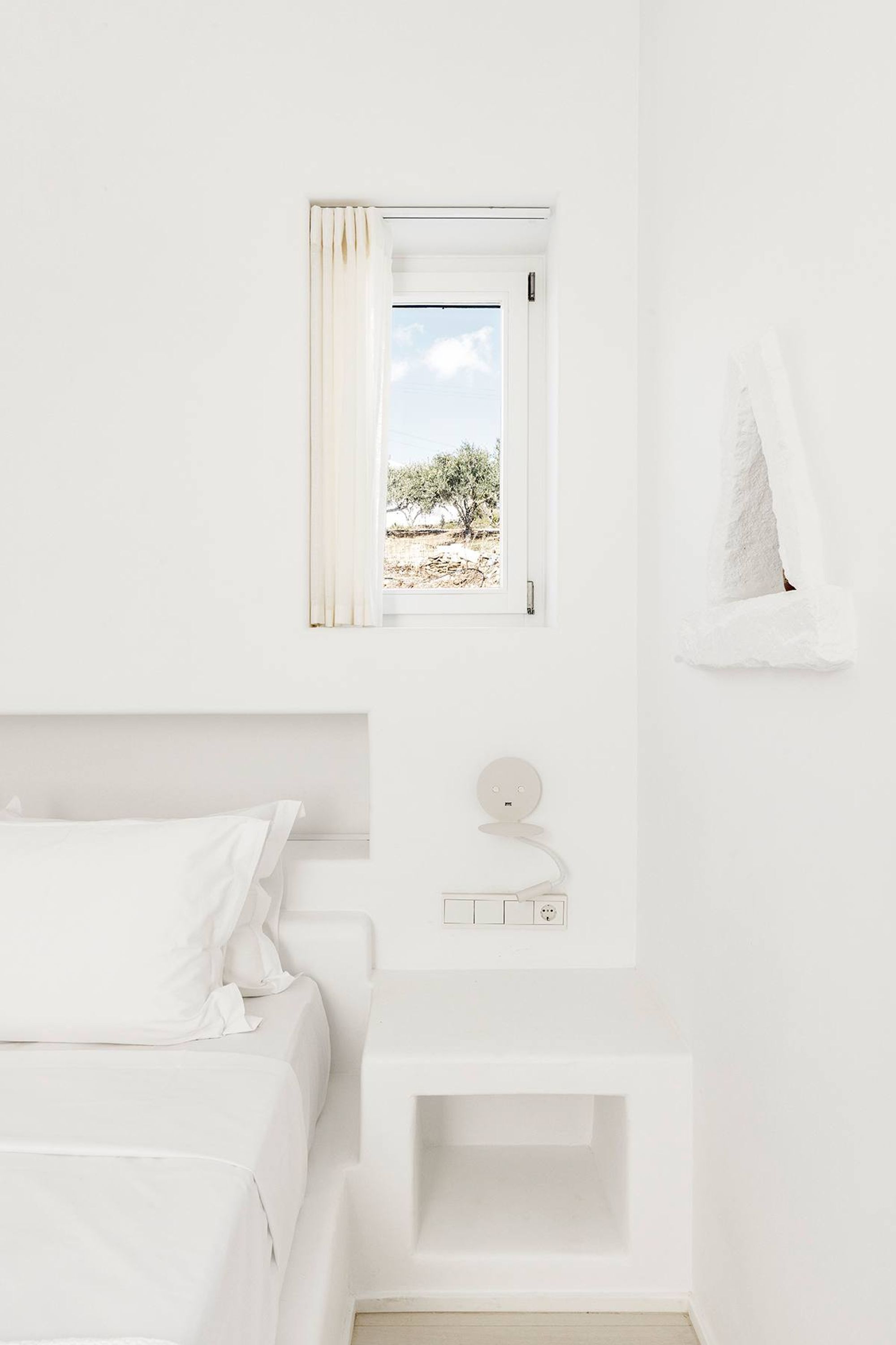 A minimalist white bedroom with a bed, bedside table, and a window with a view of a tree.