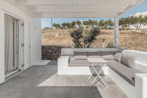 Een moderne buitenruimte met een witte bank en een kleine tafel, een stenen muur en uitzicht op een droog landschap met bomen.