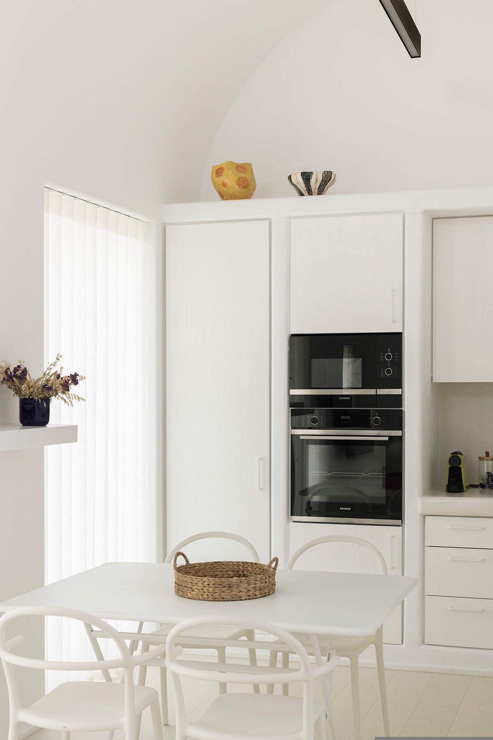Uma cozinha moderna com armários brancos, forno embutido e uma área de jantar com mesa e cadeiras brancas.