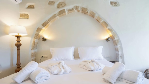 Un lit bien fait, avec du linge de lit et des serviettes blanches, une lampe de sol en bois et une arche en pierre au-dessus du lit.