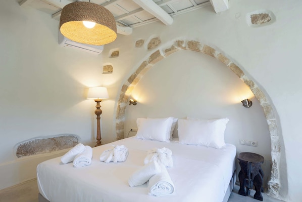 A bedroom with a large bed, white linens, and rolled towels. There is a lamp and a small wooden stool. - Aerida Guesthouse Of Stone - Natali Cozy Suite (Rethymno)