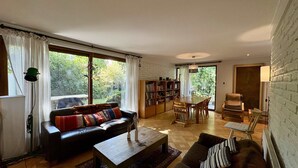 A spacious living room with a black leather sofa, a wooden coffee table, and a dining area with a table and chairs. There is a bookshelf filled with books and a large window with curtains. - House Pedro de Valdivia Norte - Garden & Parking (Las Condes)
