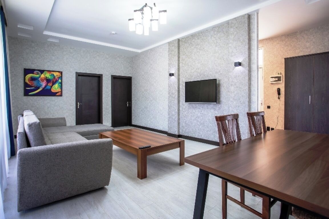 Una sala moderna con un sofá gris, una mesa de centro de madera, una mesa de comedor con sillas, un televisor montado en la pared y una puerta con un teléfono en la pared.