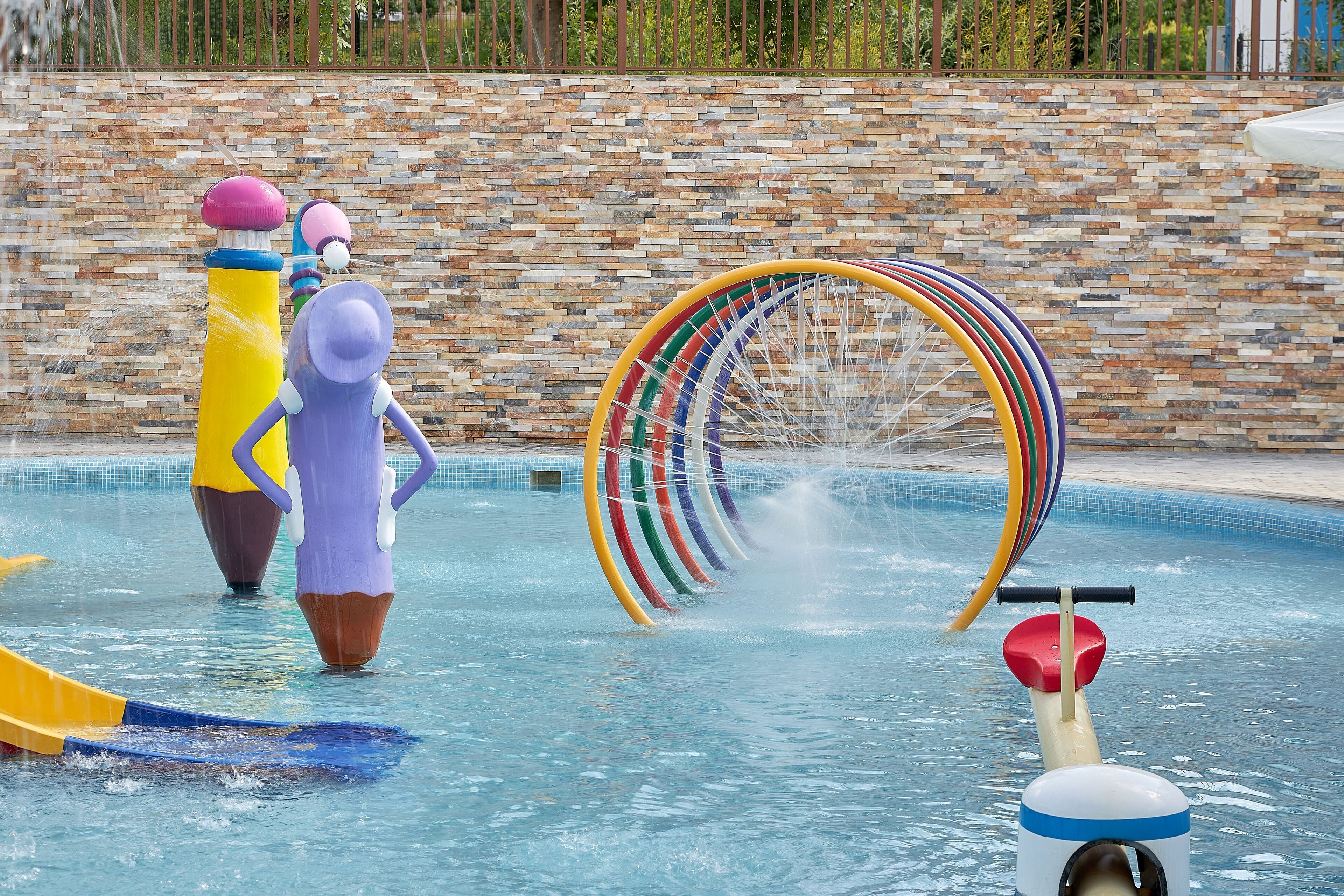 Una piscina amb tobogan aquàtic, una estructura d'anells de colors i una font d'aigua.