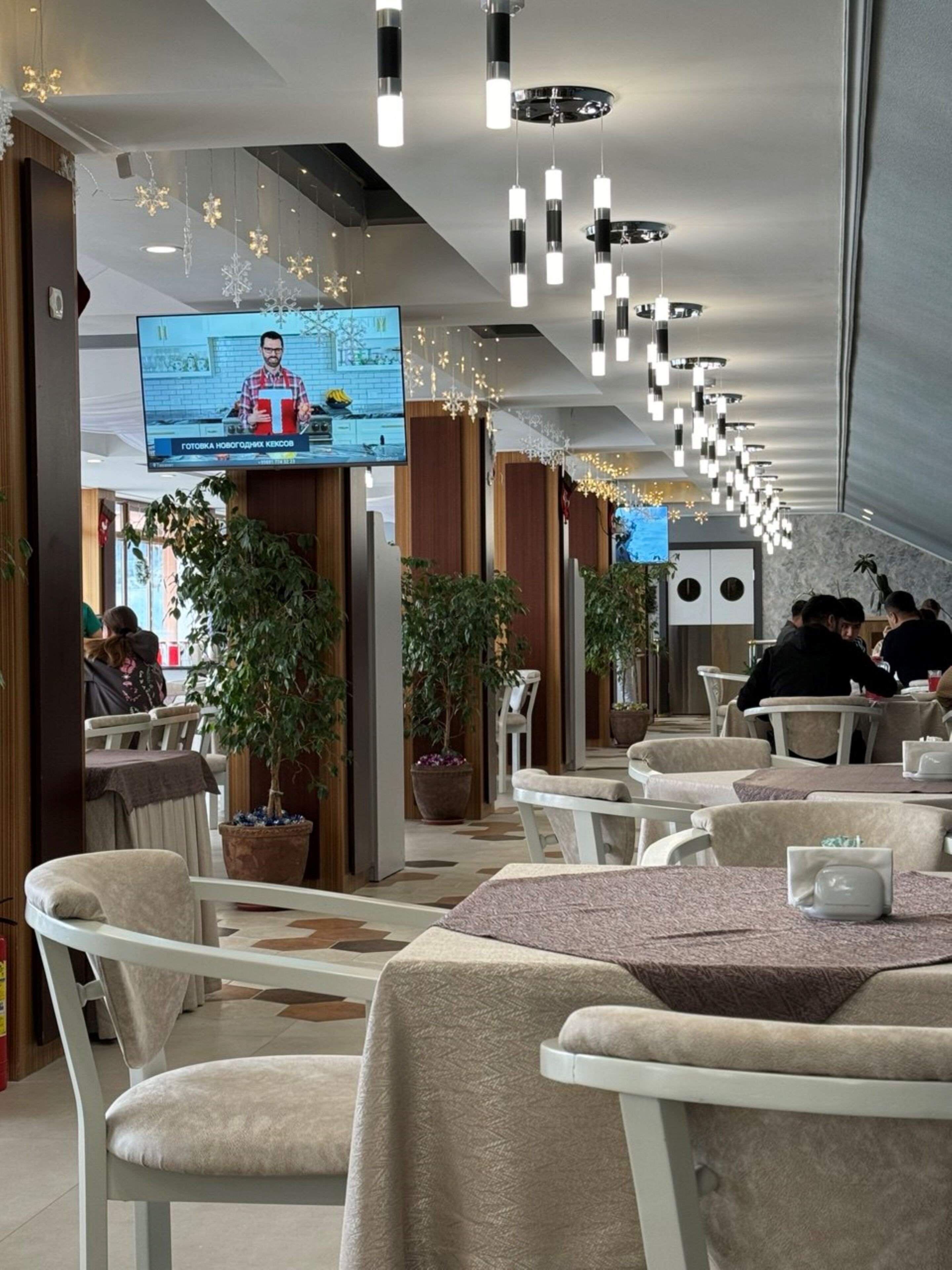 A modern restaurant with multiple tables and chairs, a television screen displaying a news broadcast, and decorative hanging lights.