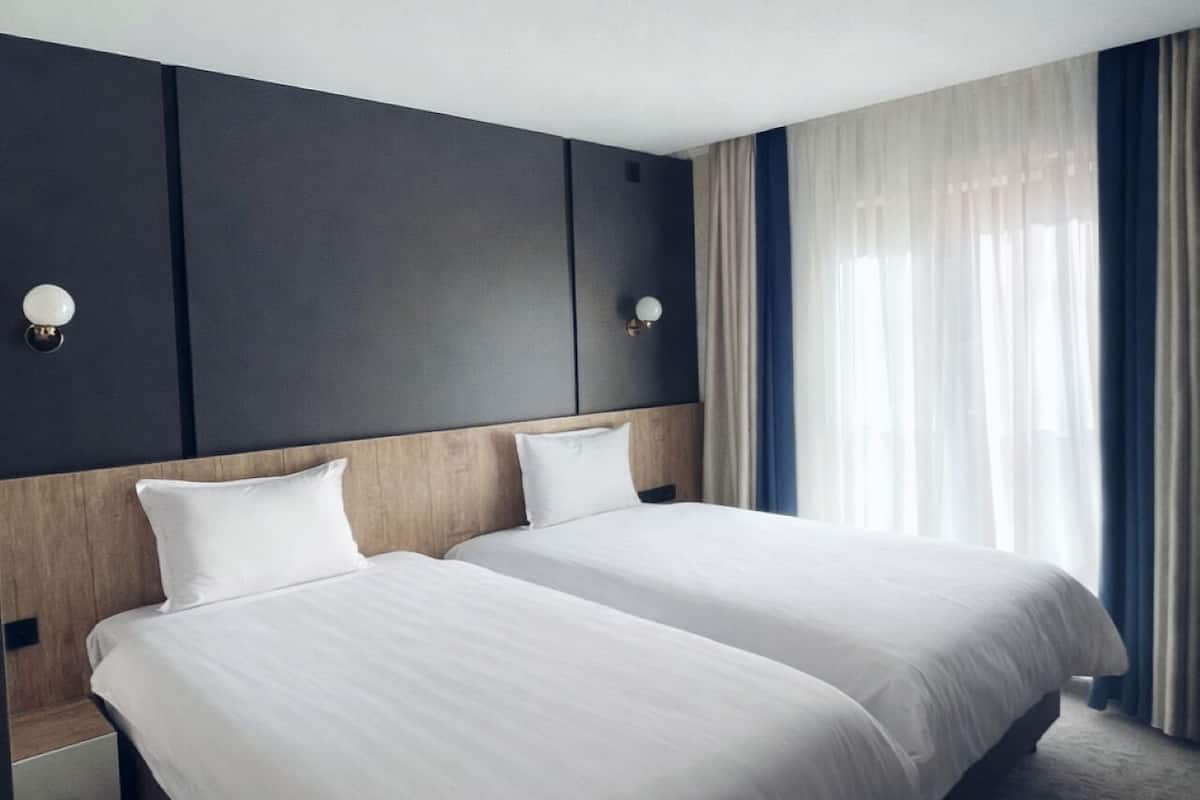 Una habitación de hotel con dos camas, un cabecero de madera, una mesita de noche y una ventana con cortinas.