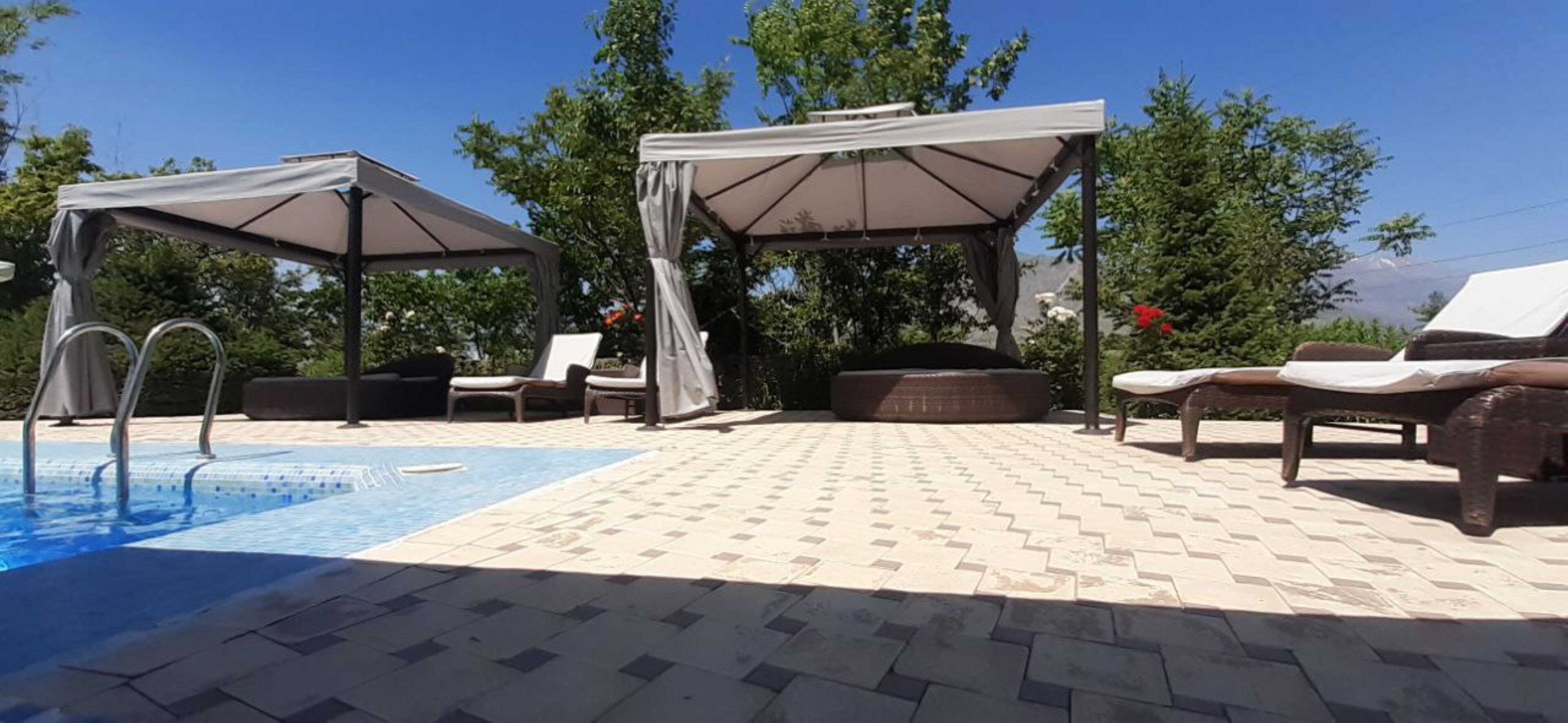 Ein Poolbereich mit Liegestühlen und Sonnenschirmen, umgeben von Bäumen und blauem Himmel.