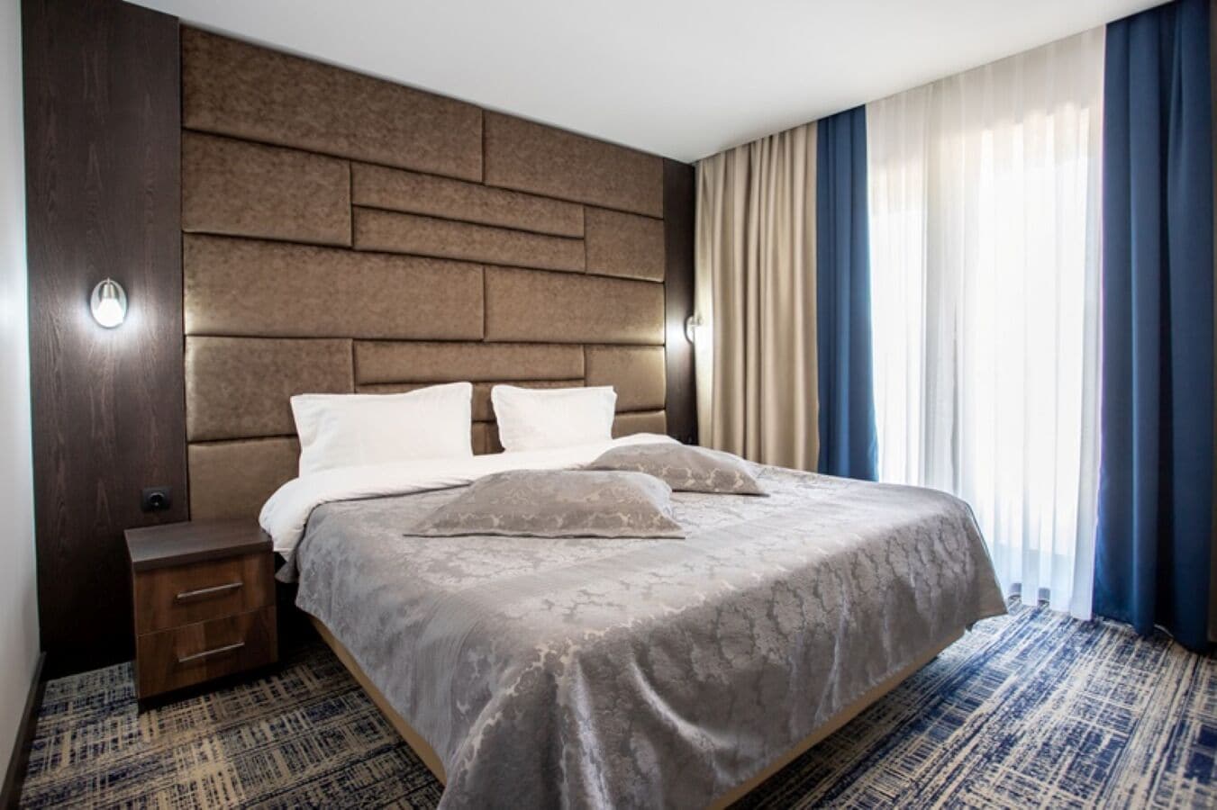 A hotel room with a large bed, a nightstand, a wall-mounted light, and a window with curtains.