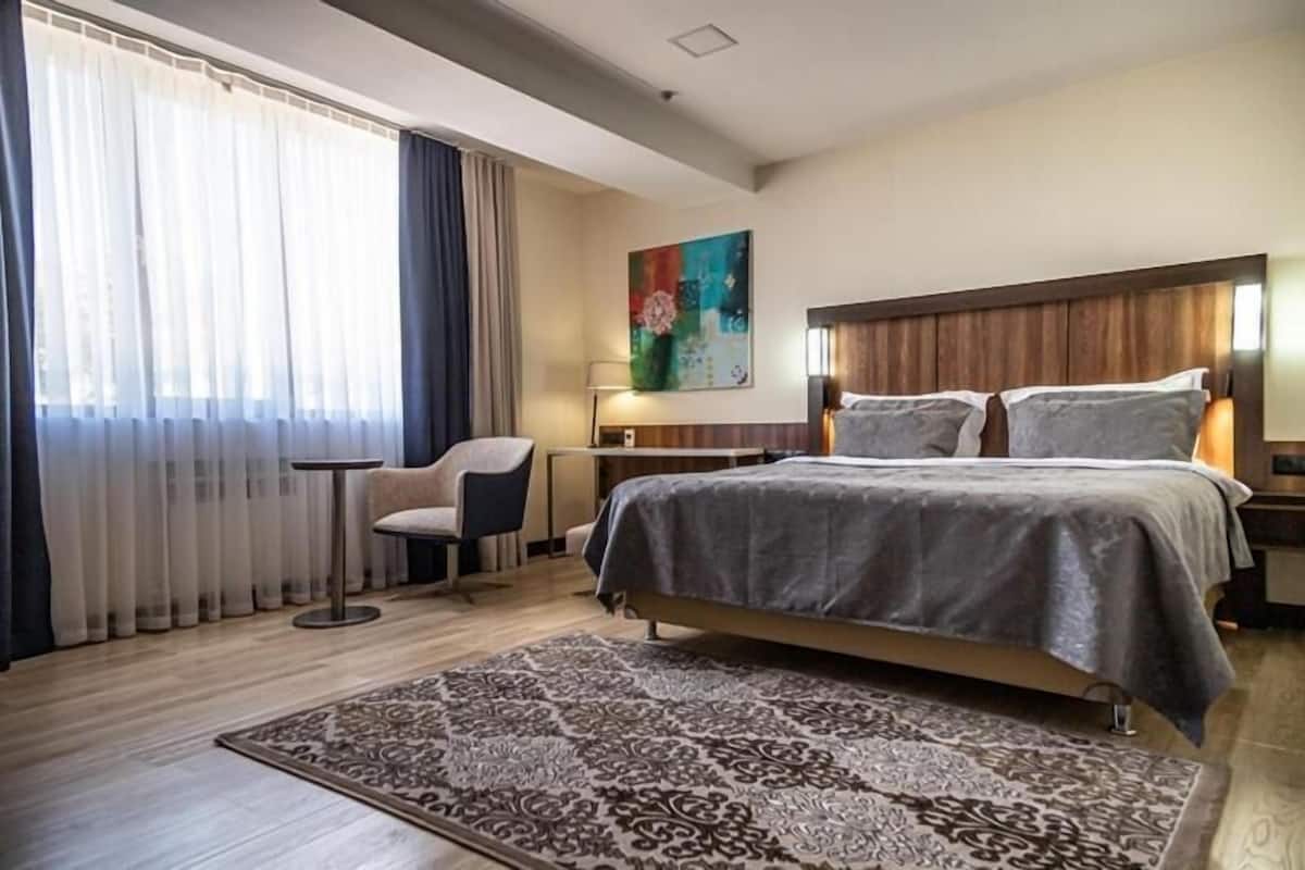 Habitación de hotel con cama, una silla, escritorio y un ventanal con cortinas.