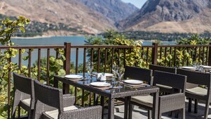 Un área de comedor al aire libre con mobiliario de mimbre y una mesa preparada para dos, con vistas a un paisaje montañoso y una masa de agua.