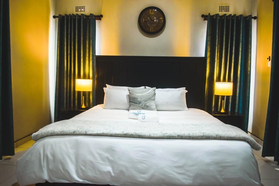 A hotel room with a large bed, two bedside lamps, a clock on the wall, and curtains on the windows.