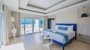 A modern hotel room with a large bed, a small side table, a decorative sculpture, and a view of the outdoors through large windows.