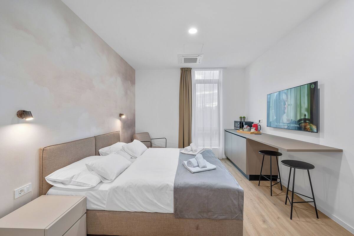 A modern hotel room with a bed, bedside tables, a desk with a chair, a TV, and a window with curtains.