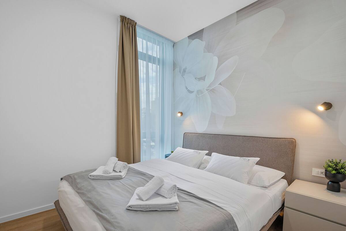 A neatly made bed with white linens and towels, a large floral wall mural, a bedside lamp, and a nightstand with a plant.