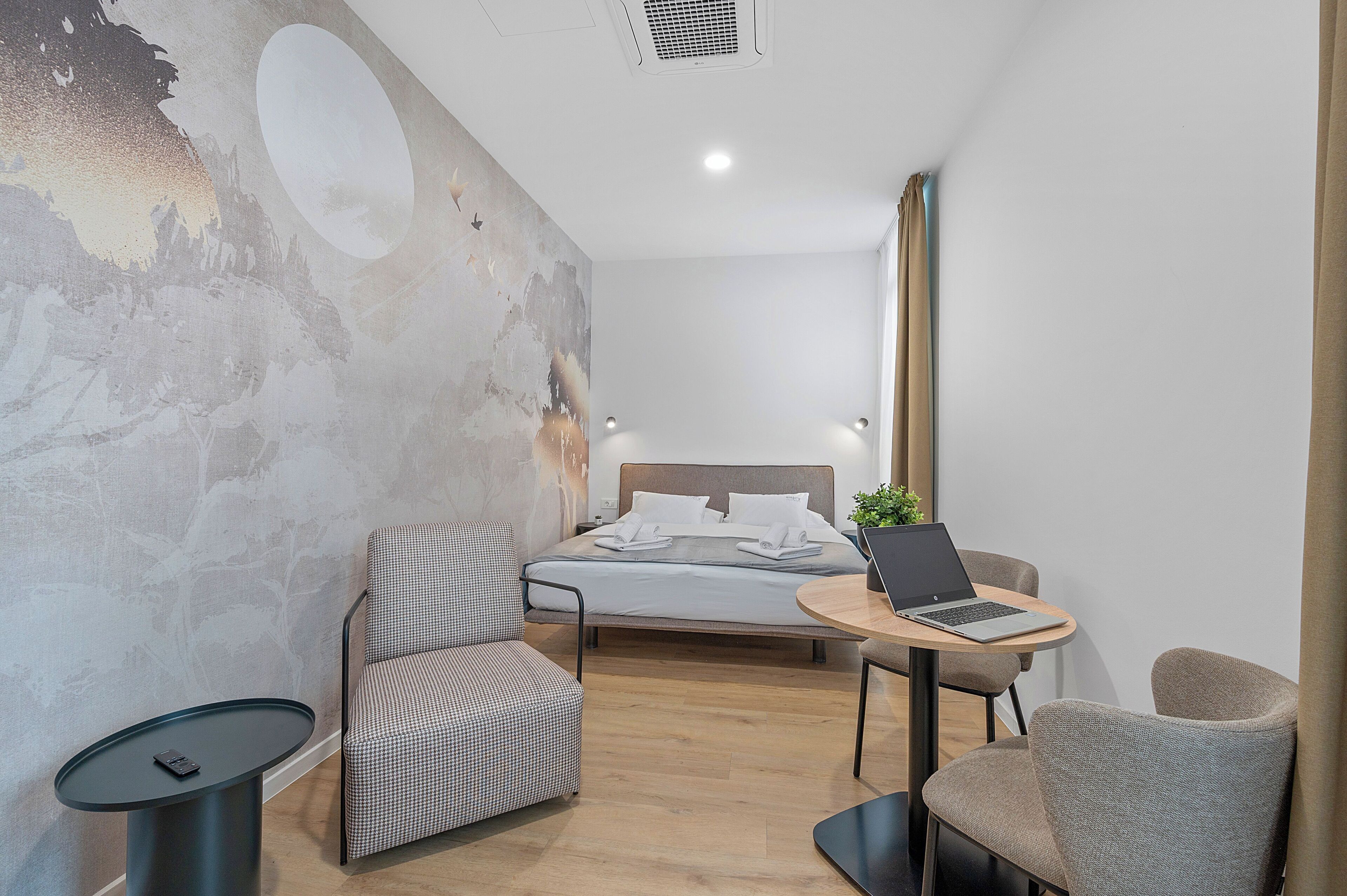 A modern hotel room with a bed, a laptop on a small table, a chair, and a round table.