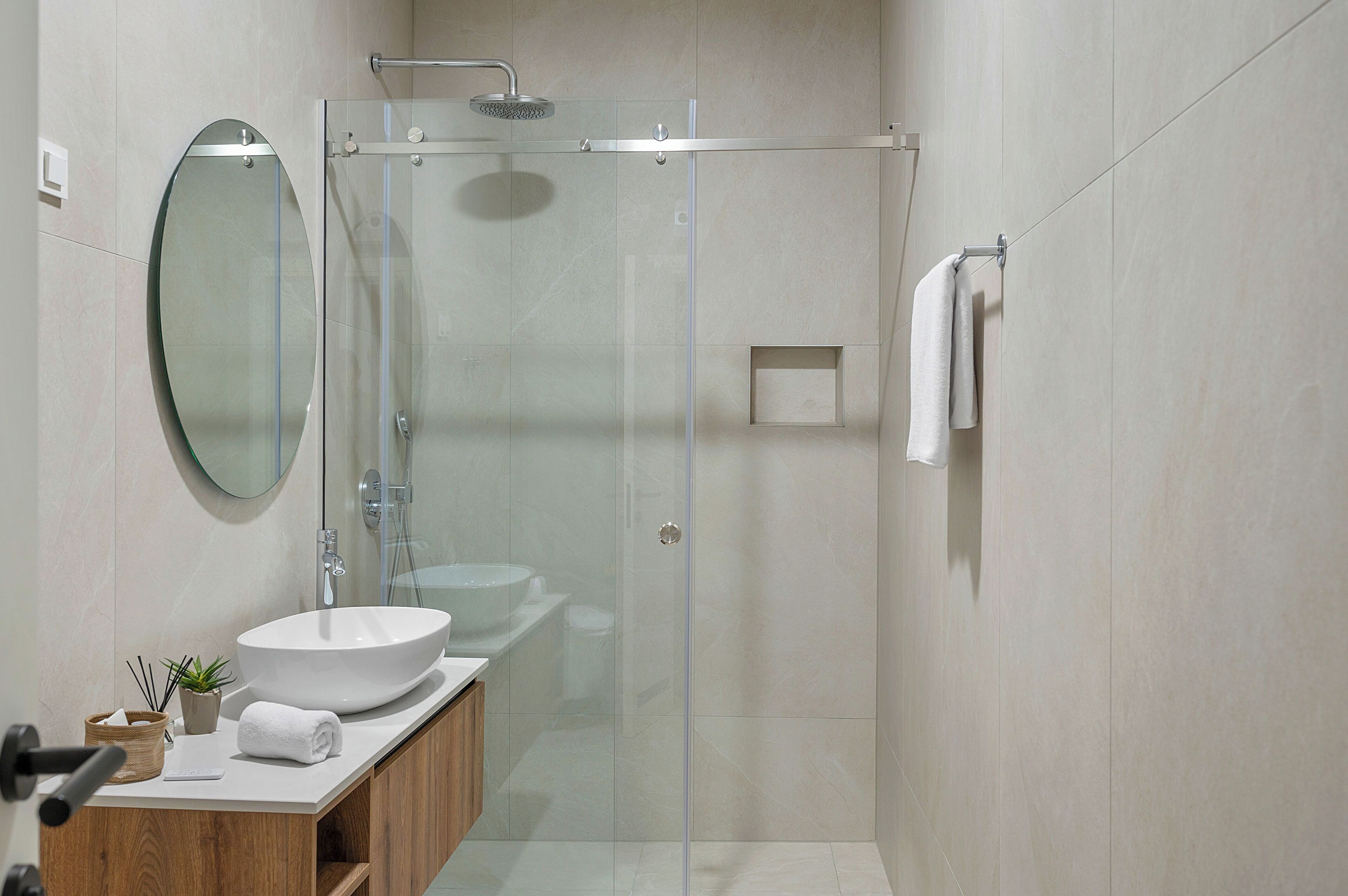 Un baño moderno con ducha envuelta en vidrio, espejo redondo, lavabo blanco y tocador de madera.