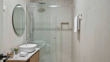 Un baño moderno con ducha envuelta en vidrio, espejo redondo, lavabo blanco y tocador de madera.