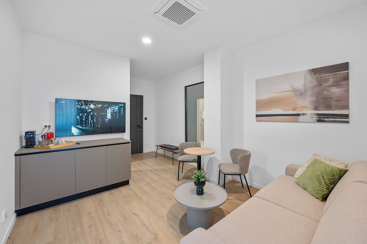 A modern living room with a flat-screen TV, a kitchen area, a dining table, and a sofa.