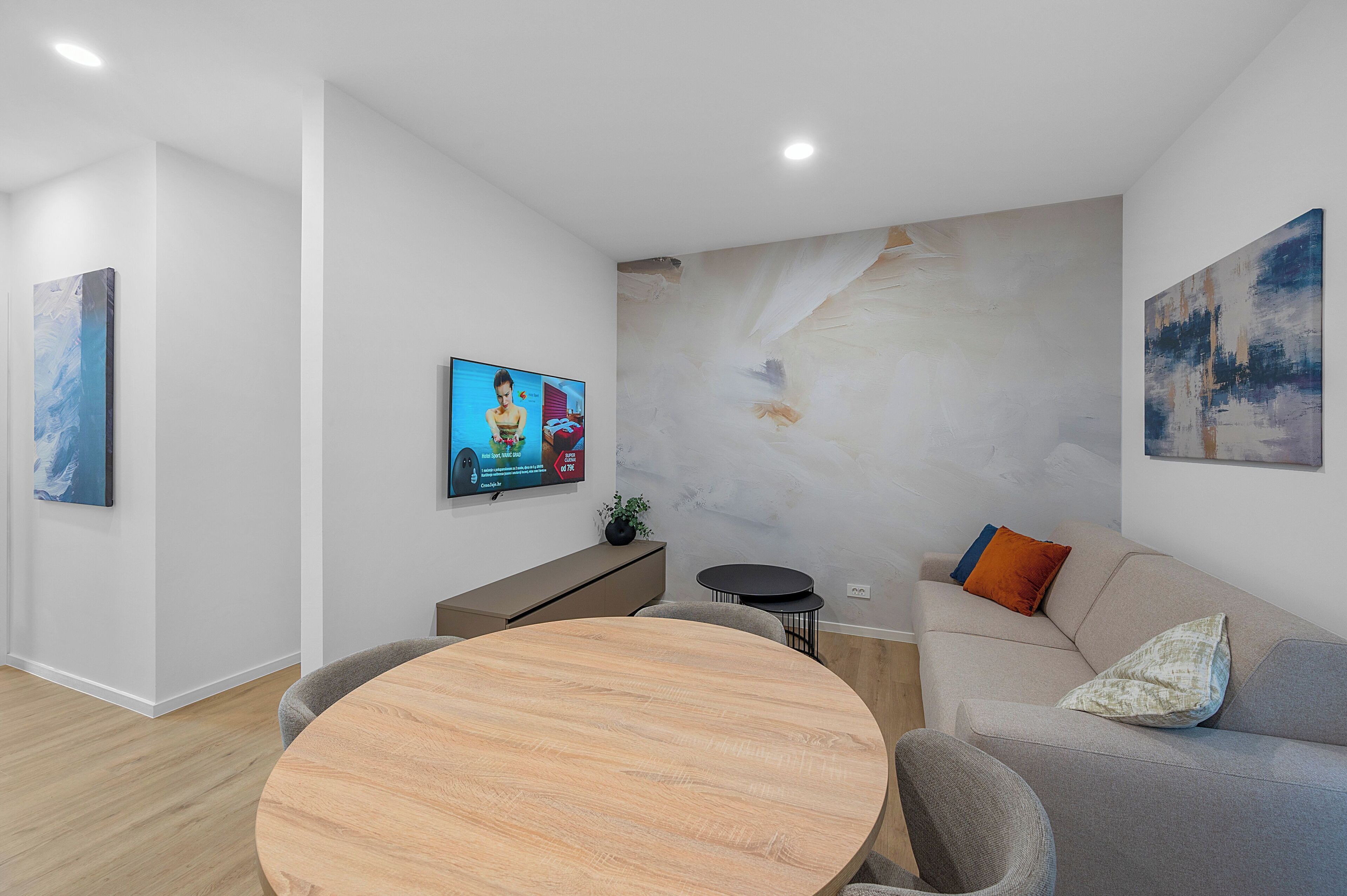 Een moderne woonkamer met een ronde houten tafel, een grijze bank met kussens, een flatscreen televisie en abstracte wanddecoratie.