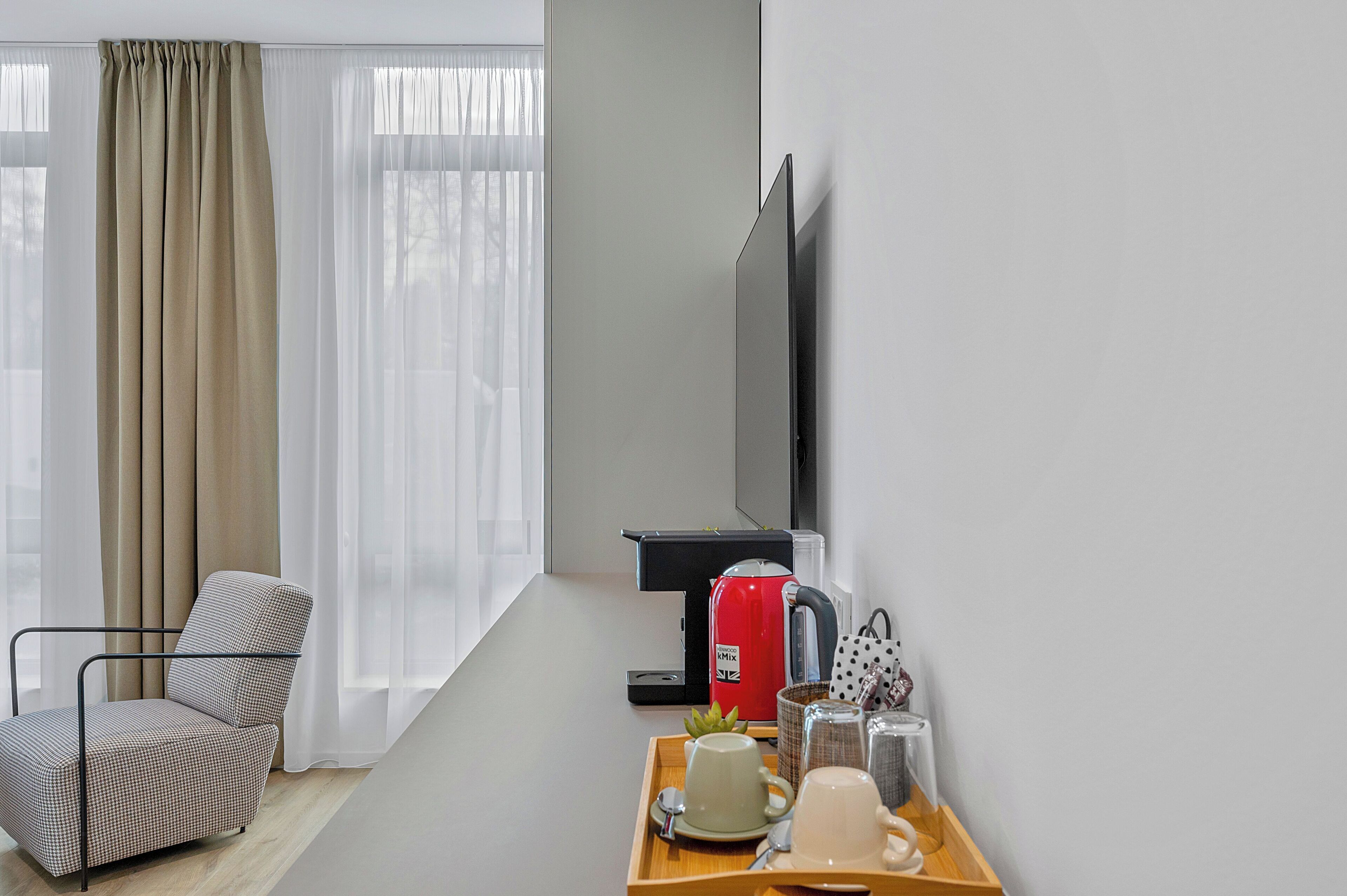 A modern hotel room with a sofa, a coffee table with a tray of breakfast items, a red kettle, and a TV.