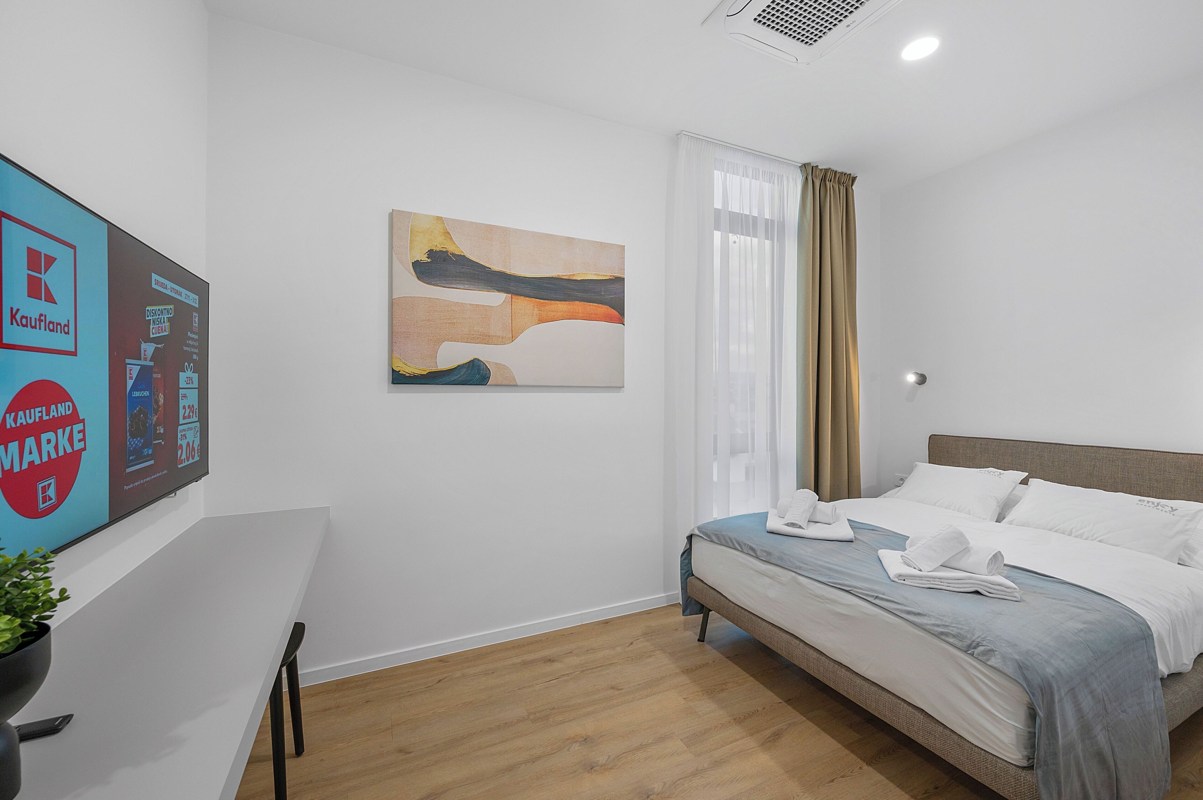 Quarto de hotel com cama, escrivaninha, planta, pintura e janela com cortinas.