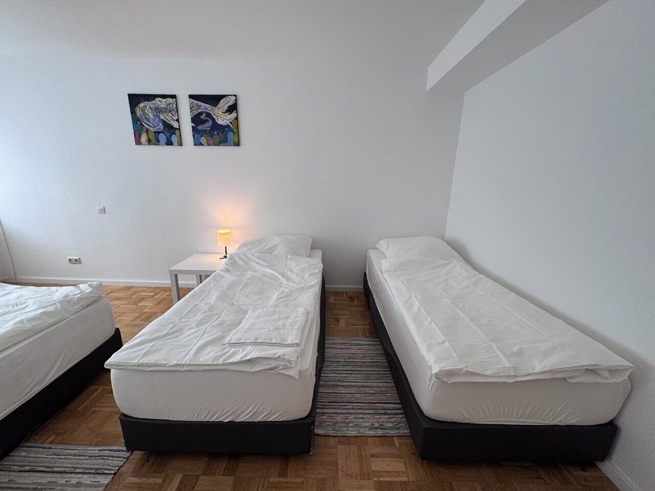 A room with three single beds, a small table with a lamp, and two paintings on the wall.