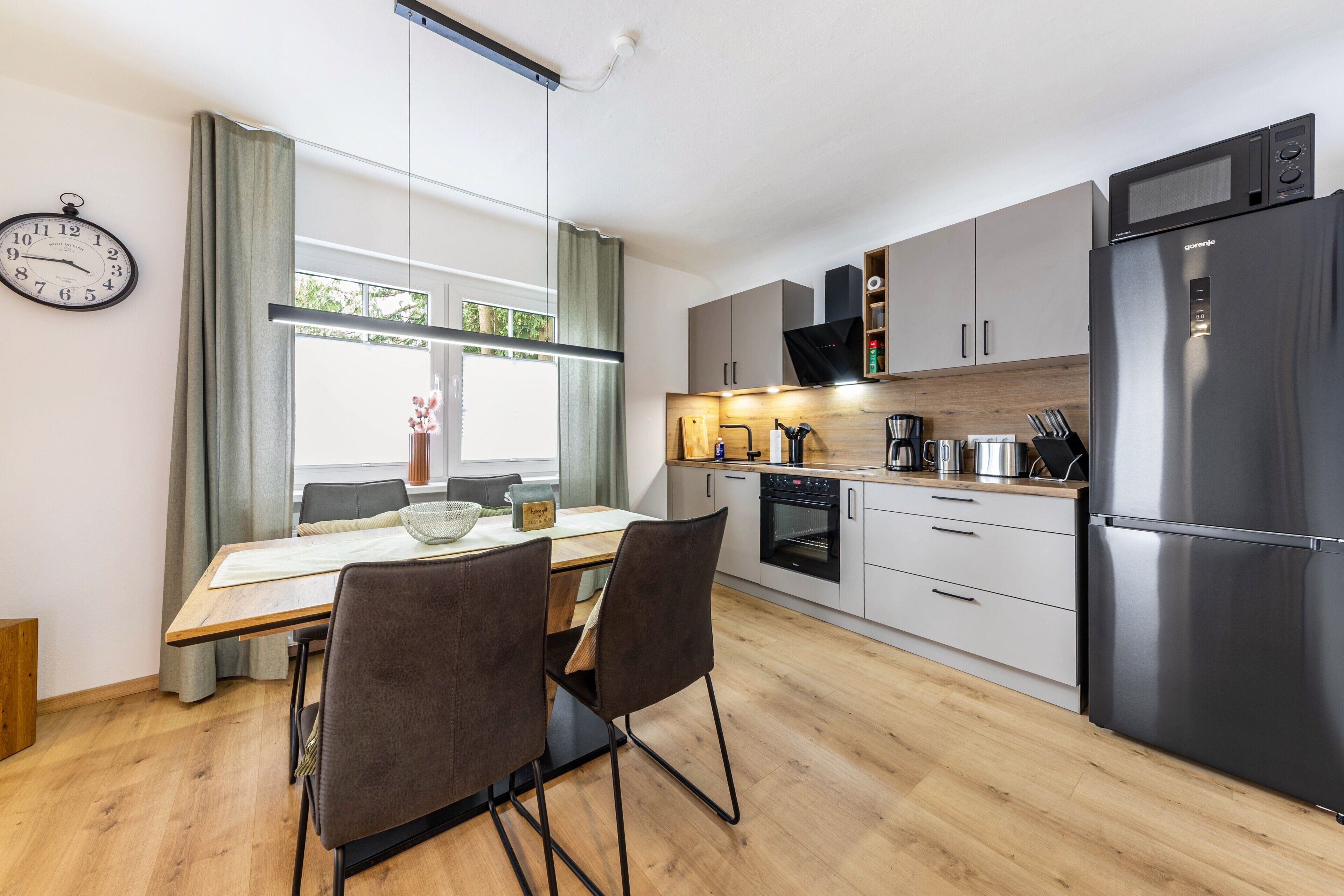 A modern kitchen with a refrigerator, oven, and microwave. There is a dining table with chairs and a clock on the wall.